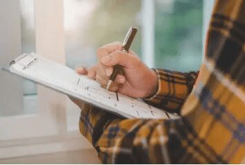 Person writing on clipboard by window.